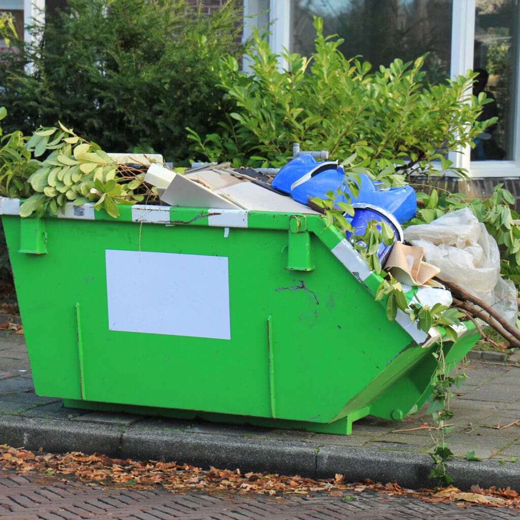 A green dumpster filled with various materials, such as branches, cardboard, and blue plastic bags, sits on a sidewalk. This scene of rubbish clearance is framed by leafy shrubs and a building facade in the background.