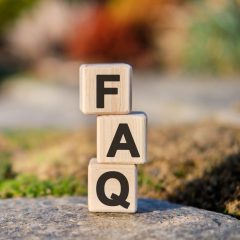 Three wooden blocks stacked vertically on a stone surface outdoors, each block displaying a letter: F on top, A in the middle, and Q at the bottom, spelling "FAQ." Blurred greenery is visible in the background.