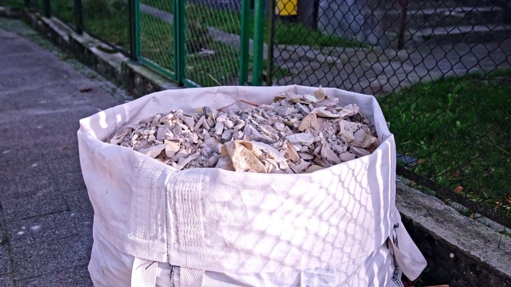 A large white fabric sack filled with broken pieces of construction debris sits on a paved pathway next to a green metal fence and grassy area.