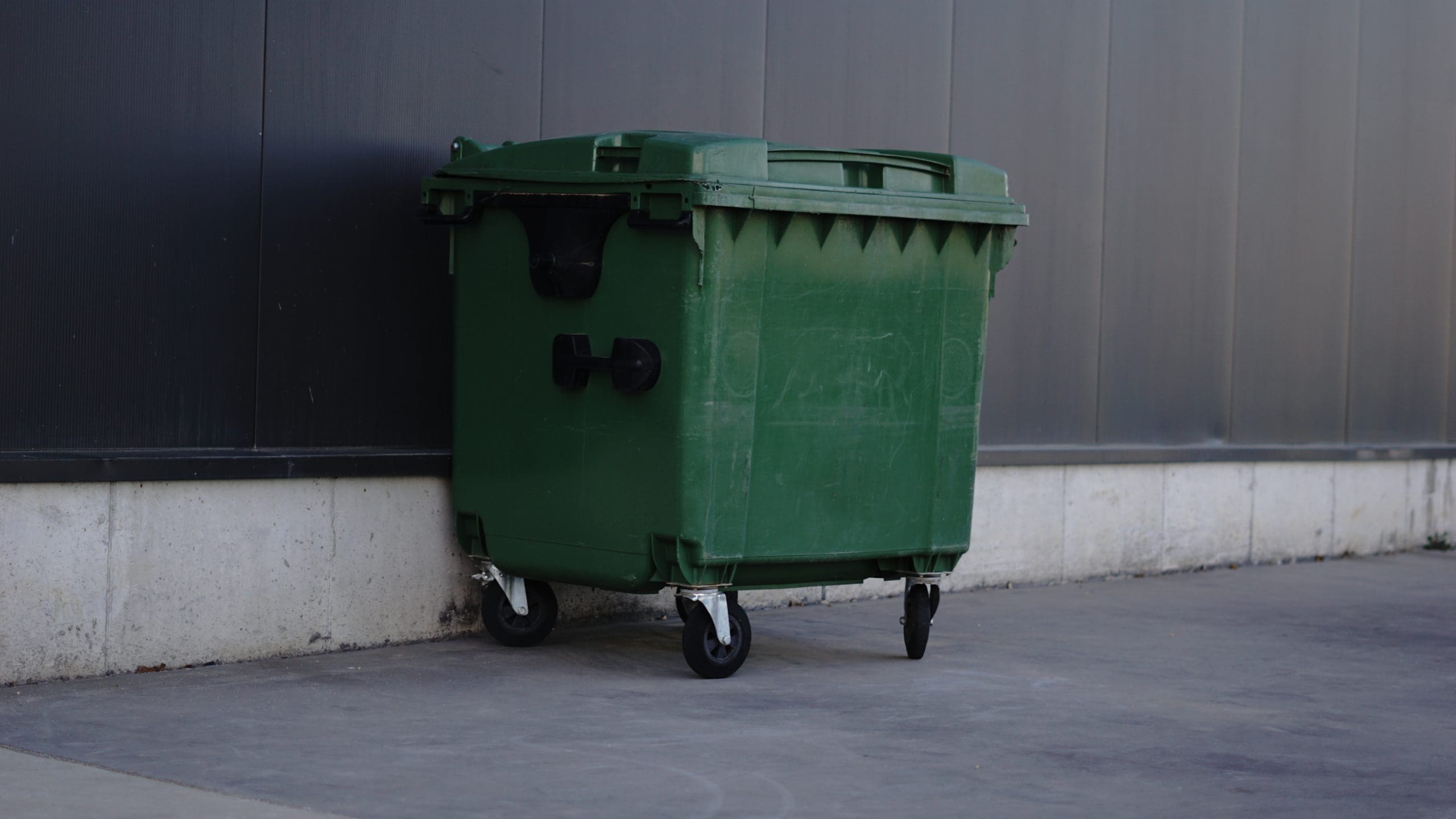 A large green outdoor garbage bin with wheels is positioned against a gray wall on a concrete pavement.
