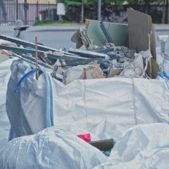Large white bags filled with construction debris, including broken drywall, metal pieces, and cardboard, are placed on a sidewalk. A bicycle and a street are visible in the background.