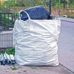 A large white industrial bag filled with materials sits on the ground near a sidewalk. Metal pieces are stacked next to the bag. A black garbage bag is partly visible inside the white bag. Fence and greenery in the background.