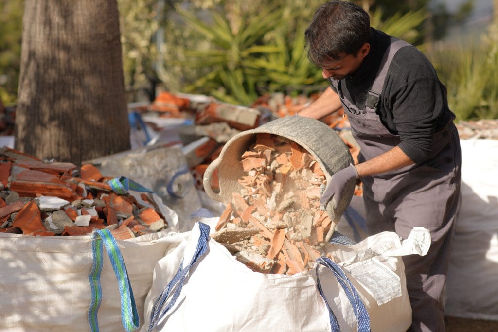 A man wearing gloves and overalls empties a bucket of construction debris, including broken bricks, into a large white bag outdoors. Other bags filled with rubble are visible in the background.